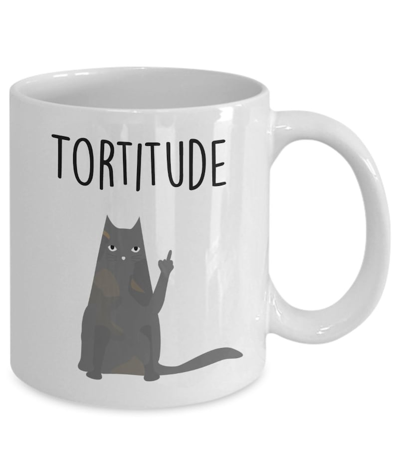 以下が含まれることがあります： 白いセラミック製のコーヒーカップ。黒い猫のイラストが中指を立てており、黒い文字で「Torttitude」と書かれている。