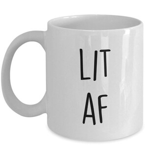 以下が含まれることがあります： 白地に黒文字で「LIT AF」と書かれた白いセラミック製のコーヒーカップです。
