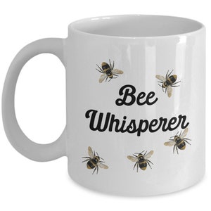 May include: White ceramic coffee mug with the text "Bee Whisperer" in black cursive font. The mug is decorated with five small images of bees.
