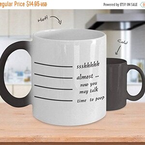 May include: A black and white color-changing mug with the text "ssshhhhh almost... now you may talk time to poop" printed on it. The mug is on a wooden surface.