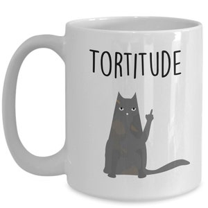 以下が含まれることがあります： 白いセラミック製のコーヒーカップ。黒い猫のイラストと「TORTITUDE」という文字が印刷されています。