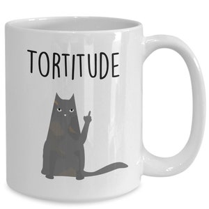 以下が含まれることがあります： 白いセラミック製のコーヒーカップ。黒い猫のイラストと「Torttitude」という文字が印刷されています。