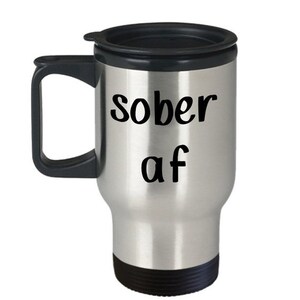 May include: A stainless steel travel mug with a black lid and handle. The mug has the text "sober af" printed on it in black.