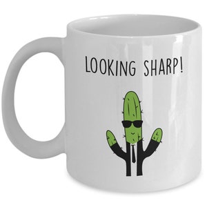 May include: White ceramic mug with a green cactus wearing sunglasses and a black tie. The text "LOOKING SHARP!" is printed on the mug.