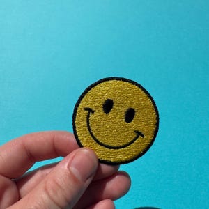 May include: A yellow smiley face patch with a black border. The patch features a smiling face with two black eyes. The patch is round and has a textured appearance. The background is a solid light blue.