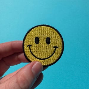 May include: Embroidered yellow smiley face patch with black detailing. The patch has a classic smiley face design with two black eyes and a curved smile. The patch is round with a black border. The background is a solid light blue.