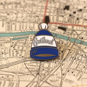 Puede incluir: Un pin de esmalte azul y blanco con forma de gorro con la palabra "Scotland" escrita en la parte delantera. El pin está sobre un mapa antiguo.