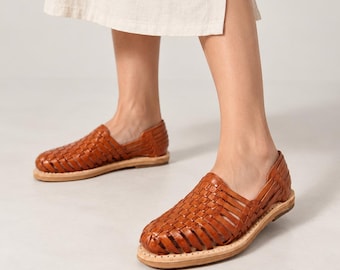 May include: A pair of brown leather huarache sandals featuring a woven lattice design across the upper, a closed rounded toe, and a flat tan sole. The sandals are worn with a light beige midi skirt that has a side slit, showing the lower legs.