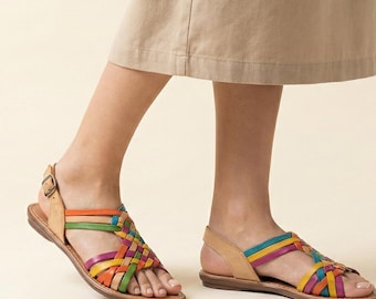 May include: Leather sandals with a colourful woven design. The sandals feature a brown sole and straps in shades of orange, green, yellow, purple, and blue. The sandals are worn by a person wearing a tan skirt.