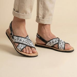 May include: Sandals with black leather straps and a white and black geometric design. The sandals have a buckle closure and a light brown sole. The person is wearing beige pants with the cuffs rolled up.