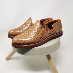 May include: A pair of tan leather woven slip-on shoes with a brown leather sole. The shoes are sitting on a small concrete stool with wooden legs.