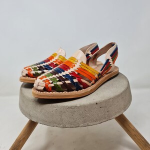 May include: A pair of colorful woven leather sandals with a slingback strap. The sandals are made of a light brown leather and have a woven pattern of red, orange, yellow, green, blue, and brown leather.