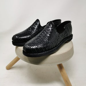 May include: A pair of black leather woven shoes. The shoes are displayed on a small, round, light gray stool with three wooden legs. The shoes have a slip-on design and a textured, woven pattern.
