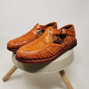 May include: A pair of handcrafted, woven leather sandals in a warm tan color. The sandals feature a closed-toe design with an open side and a buckle closure. The soles are a darker brown, and the shoes are displayed on a small, round, concrete stool.