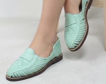 Puede incluir: Un par de zapatos planos de cuero tejido verde claro con suelas marrones. Los zapatos, con un diseño huarache clásico, se muestran en los pies de una persona, con un pie delante del otro. Son de cuero tejido.