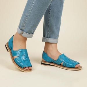 May include: Turquoise leather sandals with a woven design. The sandals have a flat sole with a light brown base and a black and white stripe. The sandals are worn with light blue denim jeans.