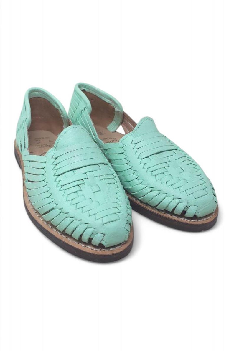 May include: A pair of mint green huarache sandals. The sandals have a woven leather design on the upper and a brown sole. The open-toe shoes are displayed on a white background. The sandals are a type of footwear.