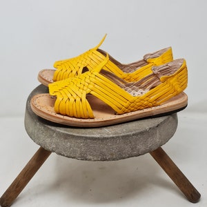 May include: A pair of yellow leather sandals with woven straps. The sandals are sitting on a wooden stool with a stone top.