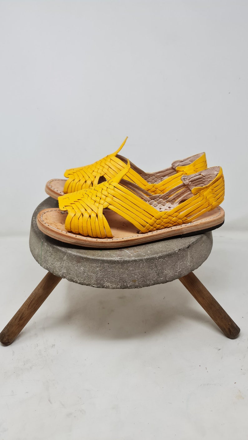 May include: A pair of yellow leather sandals with woven straps. The sandals are sitting on a wooden stool with a stone top.