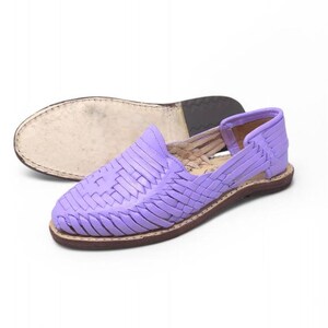 May include: A pair of lavender woven leather huarache sandals. The sandals feature a closed toe design with a woven vamp and a brown sole. The second sandal is upside down, showing the sole. The shoes are a light purple color.