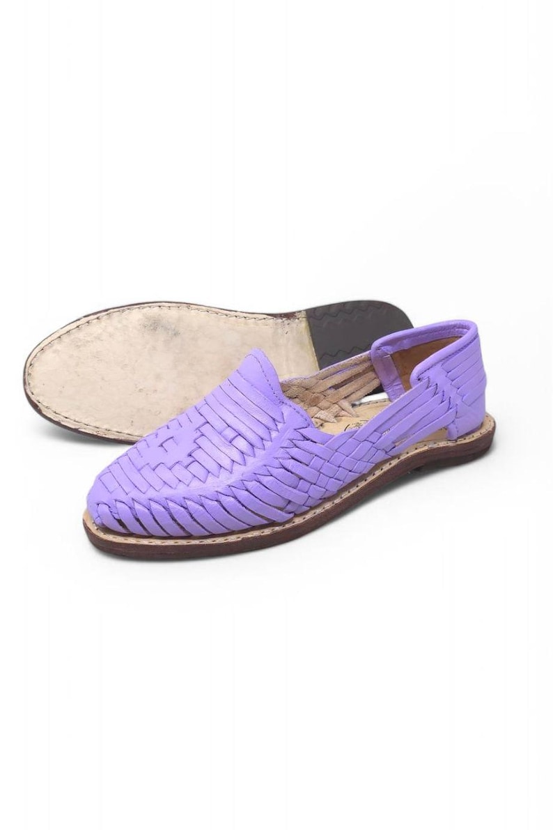 May include: A pair of lavender woven leather huarache sandals. The sandals feature a closed toe design with a woven vamp and a brown sole. The second sandal is upside down, showing the sole. The shoes are a light purple color.