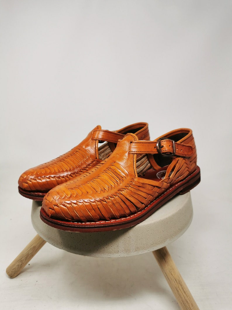 May include: A pair of handcrafted, woven leather sandals in a warm tan color. The sandals feature a closed-toe design with an open side and a buckle closure. The soles are a darker brown, and the shoes are displayed on a small, round, concrete stool.