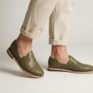 May include: Olive green woven leather slip-on shoes. The shoes have a rounded toe and a low profile, with a woven design across the top. The sole is a light tan color with a thin red stripe and a black stripe.