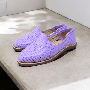 May include: A pair of lavender woven leather shoes. The shoes have a slip-on design with an open heel and a brown sole. The woven pattern covers the top of the shoe. The shoes are displayed on a light gray surface.