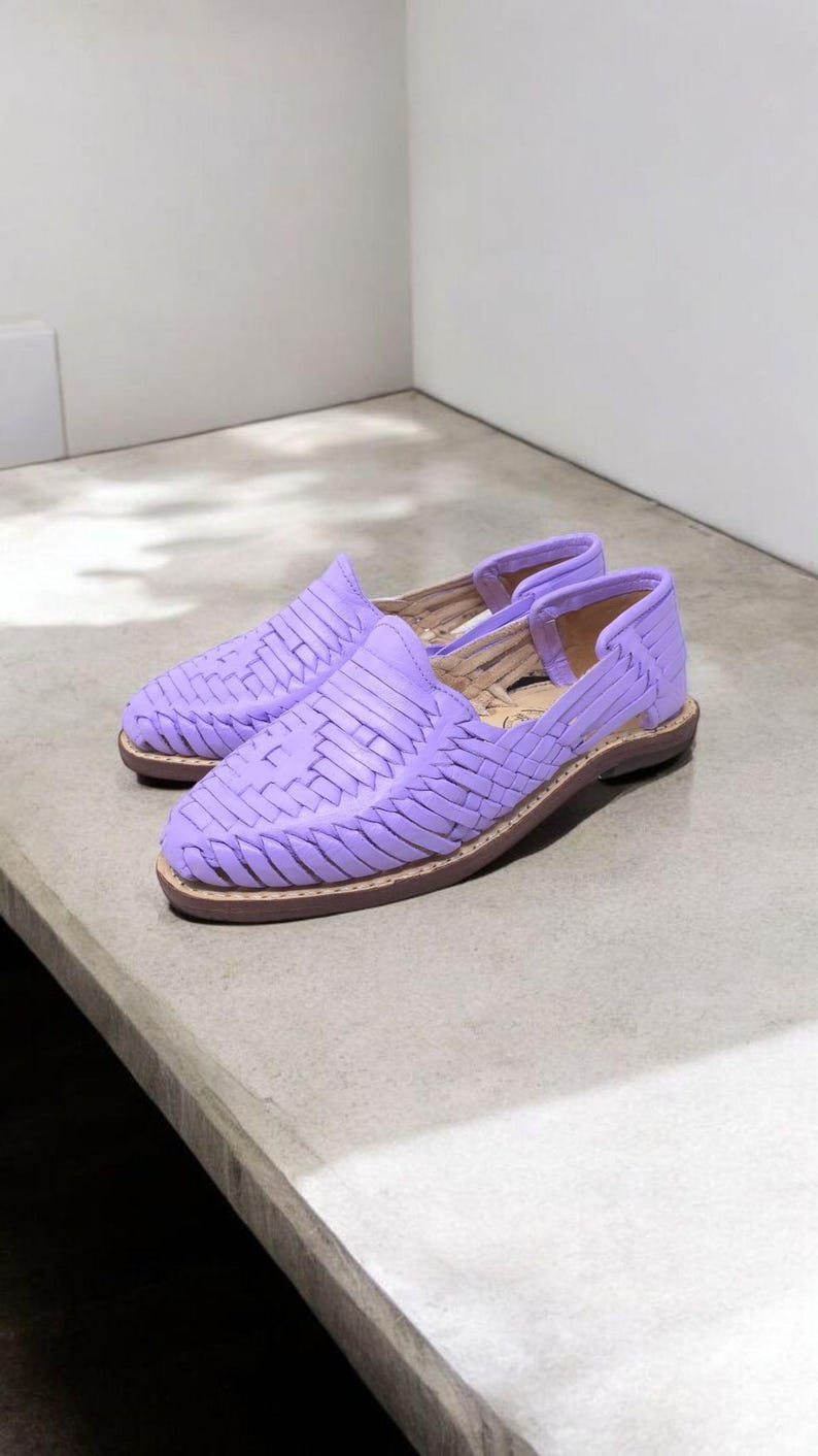 May include: A pair of lavender woven leather shoes. The shoes have a slip-on design with an open heel and a brown sole. The woven pattern covers the top of the shoe. The shoes are displayed on a light gray surface.