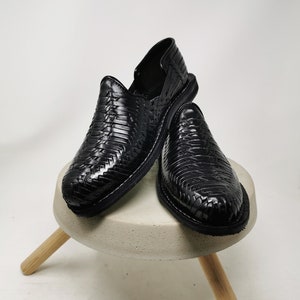 May include: A pair of black leather woven shoes. The shoes are displayed on a small, round, light gray concrete stool with three wooden legs. The shoes have a slip-on design and a textured, basket-weave pattern.