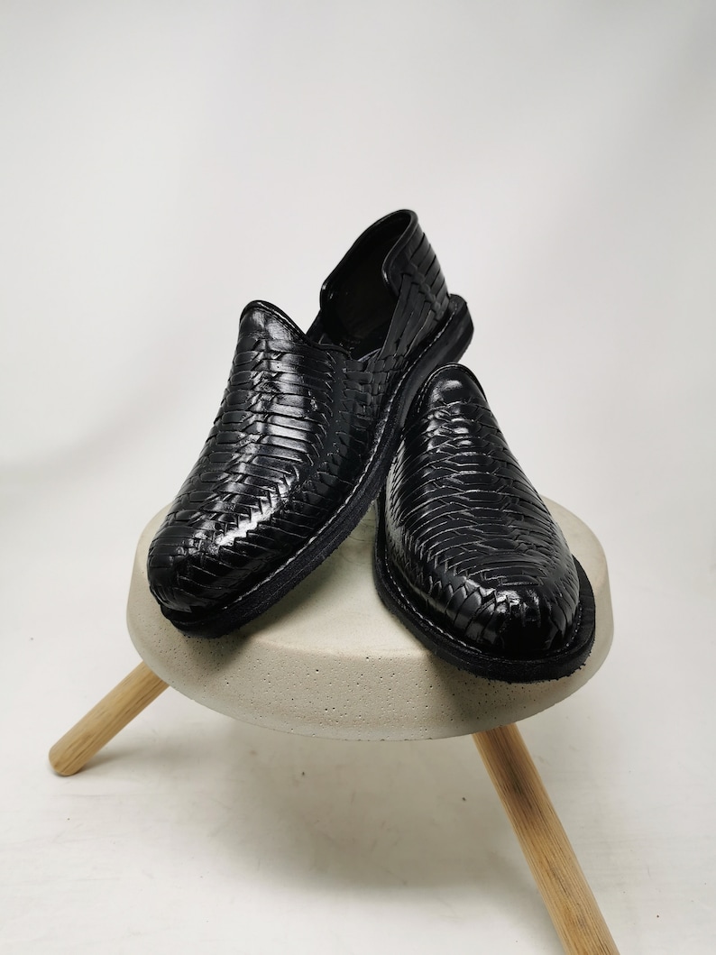 May include: A pair of black leather woven shoes. The shoes are displayed on a small, round, light gray concrete stool with three wooden legs. The shoes have a slip-on design and a textured, basket-weave pattern.