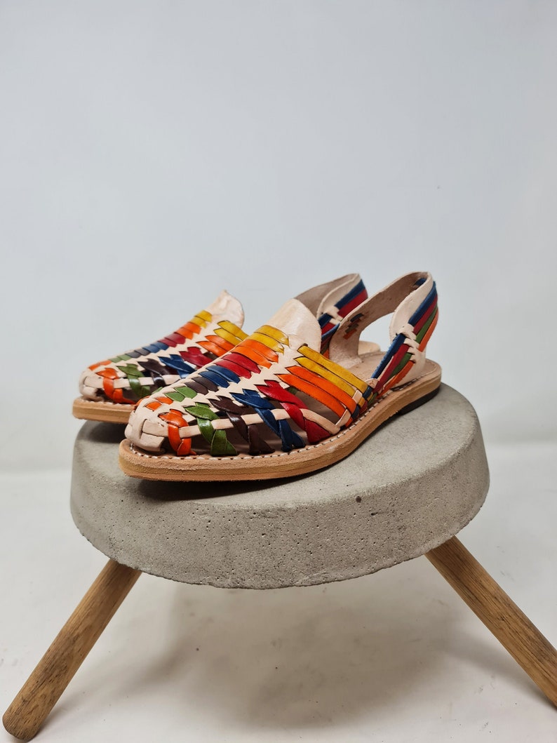 May include: A pair of colorful woven leather sandals with a slingback strap. The sandals are made of a light brown leather and have a woven pattern of red, orange, yellow, green, blue, and brown leather.