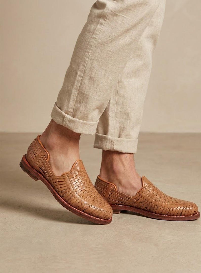May include: Tan leather huarache shoes featuring a woven upper design with a closed toe and an open heel section. The shoes have a low stacked heel and a dark brown sole, paired with light beige cuffed trousers.