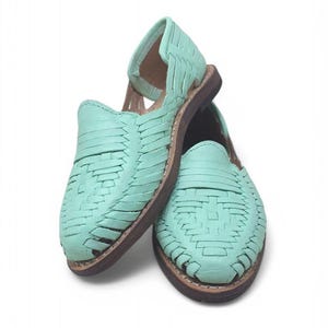 May include: A pair of mint green huarache sandals. The sandals have a woven leather design and a brown sole. The open-toe shoes are displayed on a white background. These sandals are a summer footwear option.