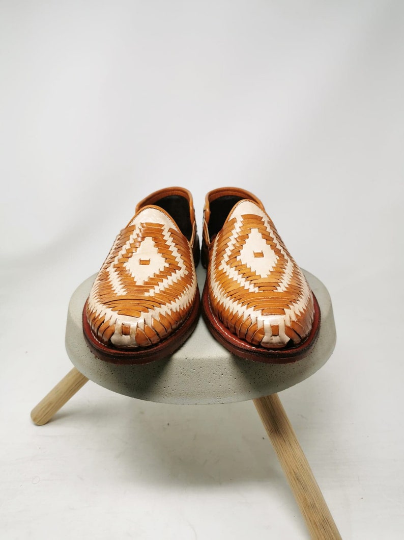 K&ouml;nnte beinhalten: Paar gewebte Loafer aus hellbraunem und cremefarbenem Leder. Die Schuhe haben ein Rautenmuster und eine braune Ledersohle. Sie werden auf einem kleinen, grauen Betonhocker mit Holzbeinen pr&auml;sentiert.