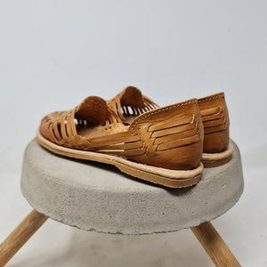 Puede incluir: Sandalias de cuero marr&oacute;n claro con dise&ntilde;o tejido. Se exhiben sobre un peque&ntilde;o taburete redondo de hormig&oacute;n gris con patas de madera. Las sandalias tienen suela plana.