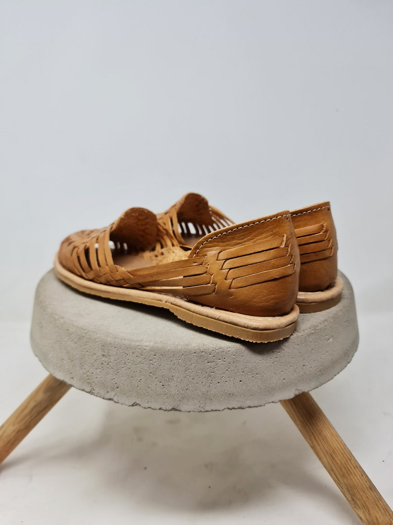 Puede incluir: Sandalias de cuero marr&oacute;n claro con dise&ntilde;o tejido. Se exhiben sobre un peque&ntilde;o taburete redondo de hormig&oacute;n gris con patas de madera. Las sandalias tienen suela plana.