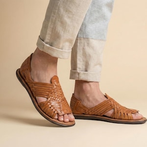 May include: Brown leather sandals with a woven design. The sandals have a flat sole and a slip-on style. The image shows the sandals on a person's feet, with the sandals in focus. The sandals are a warm brown color.