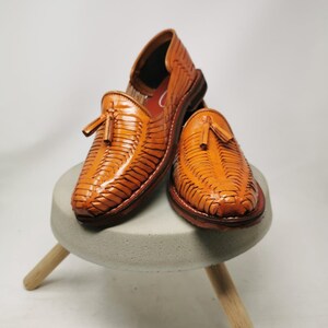 Puede incluir: Un par de sandalias de cuero marr&oacute;n con borlas. Las sandalias son tejidas y tienen una punta puntiaguda. Est&aacute;n sentadas en un taburete de madera.