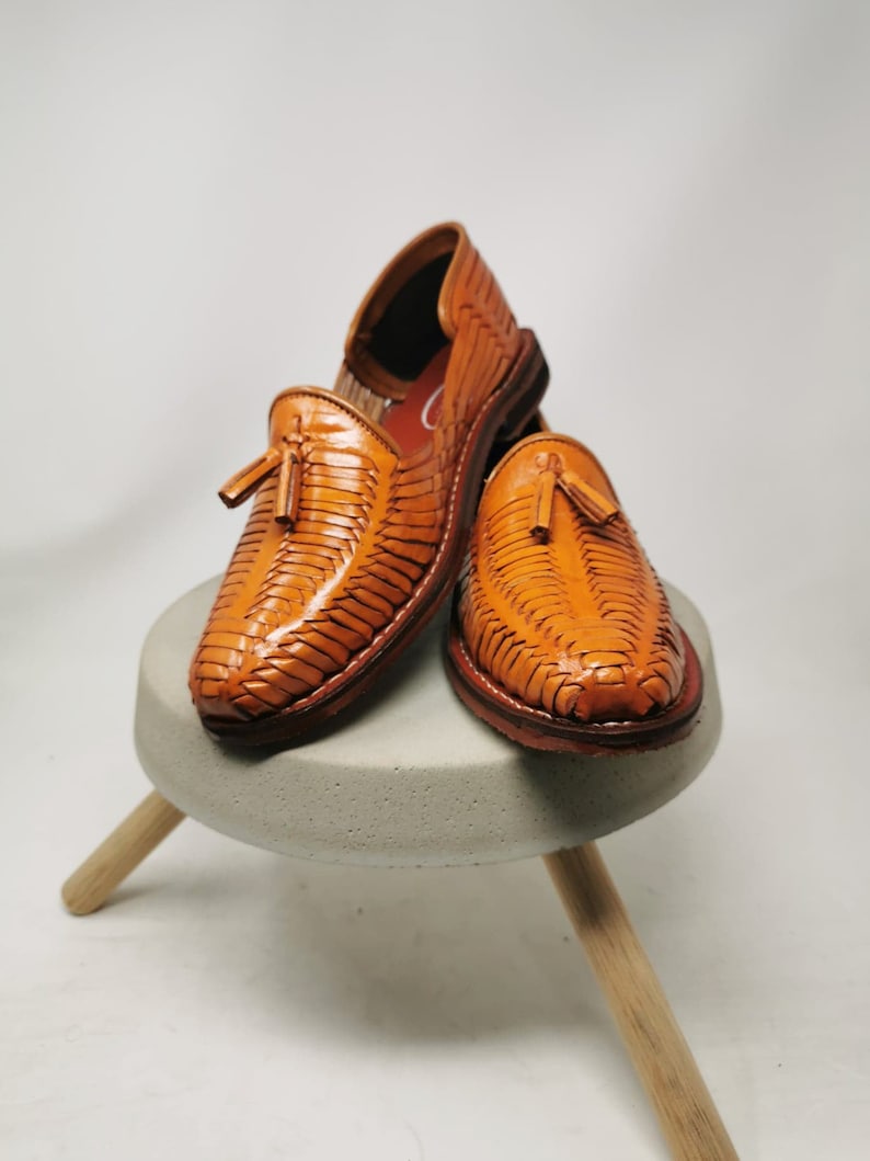 Puede incluir: Un par de sandalias de cuero marr&oacute;n con borlas. Las sandalias son tejidas y tienen una punta puntiaguda. Est&aacute;n sentadas en un taburete de madera.