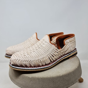 Mocasines trenzados en tono crema, con ribete colorido y suela de esparto, reposan sobre un pedestal de concreto, evocando un estilo minimalista y moderno. Perfectos para resaltar la moda sostenible.