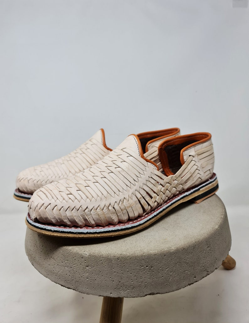 Mocasines trenzados en tono crema, con ribete colorido y suela de esparto, reposan sobre un pedestal de concreto, evocando un estilo minimalista y moderno. Perfectos para resaltar la moda sostenible.