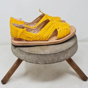 May include: A pair of yellow leather sandals with a woven design. The sandals are sitting on a wooden stool.