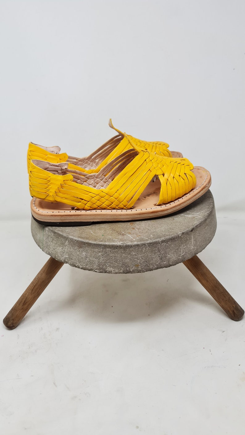 May include: A pair of yellow leather sandals with a woven design. The sandals are sitting on a wooden stool.