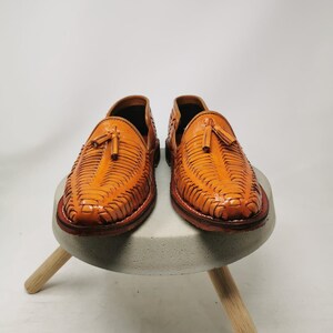 Puede incluir: Un par de sandalias de cuero marr&oacute;n con un dise&ntilde;o tejido y borlas. Las sandalias est&aacute;n sobre un taburete de hormig&oacute;n con patas de madera.