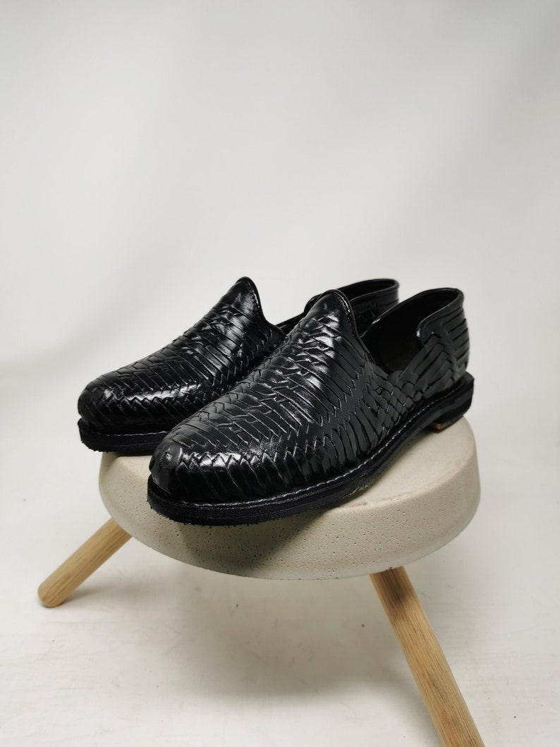 May include: A pair of black leather woven shoes. The shoes are displayed on a small, round, light gray stool with three wooden legs. The shoes have a slip-on design and a textured, woven pattern.