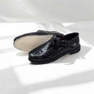 May include: Black leather huaraches sandals with woven design and buckle closure. One sandal is upright, the other is upside down, showing the sole. The sandals are on a white background.