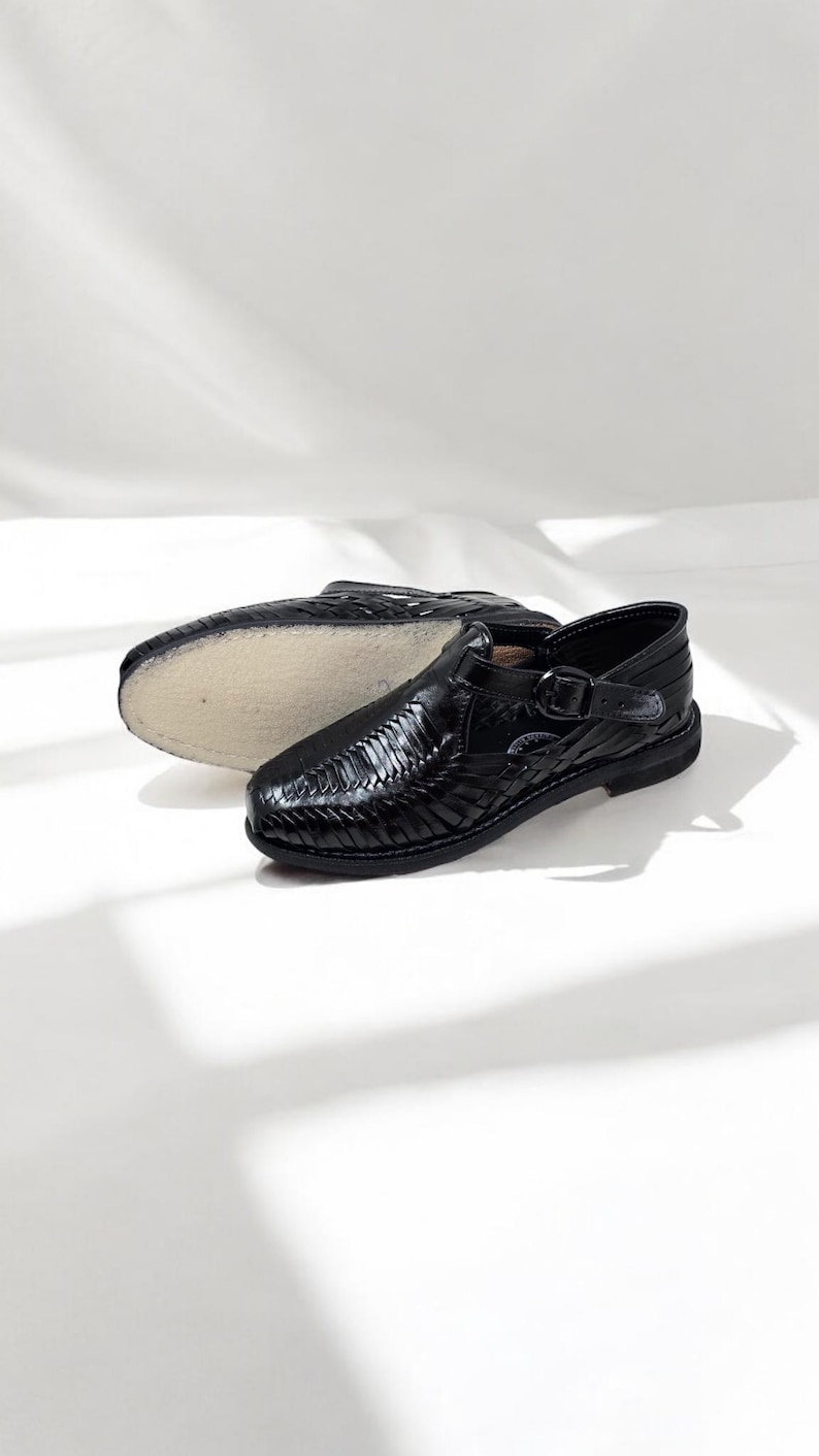 May include: Black leather huaraches sandals with woven design and buckle closure. One sandal is upright, the other is upside down, showing the sole. The sandals are on a white background.