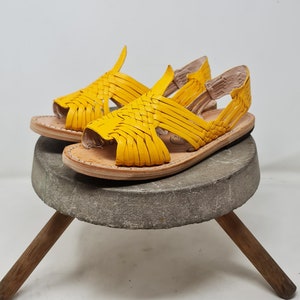 May include: A pair of yellow leather sandals with woven straps. The sandals are sitting on a wooden stool with a stone top.