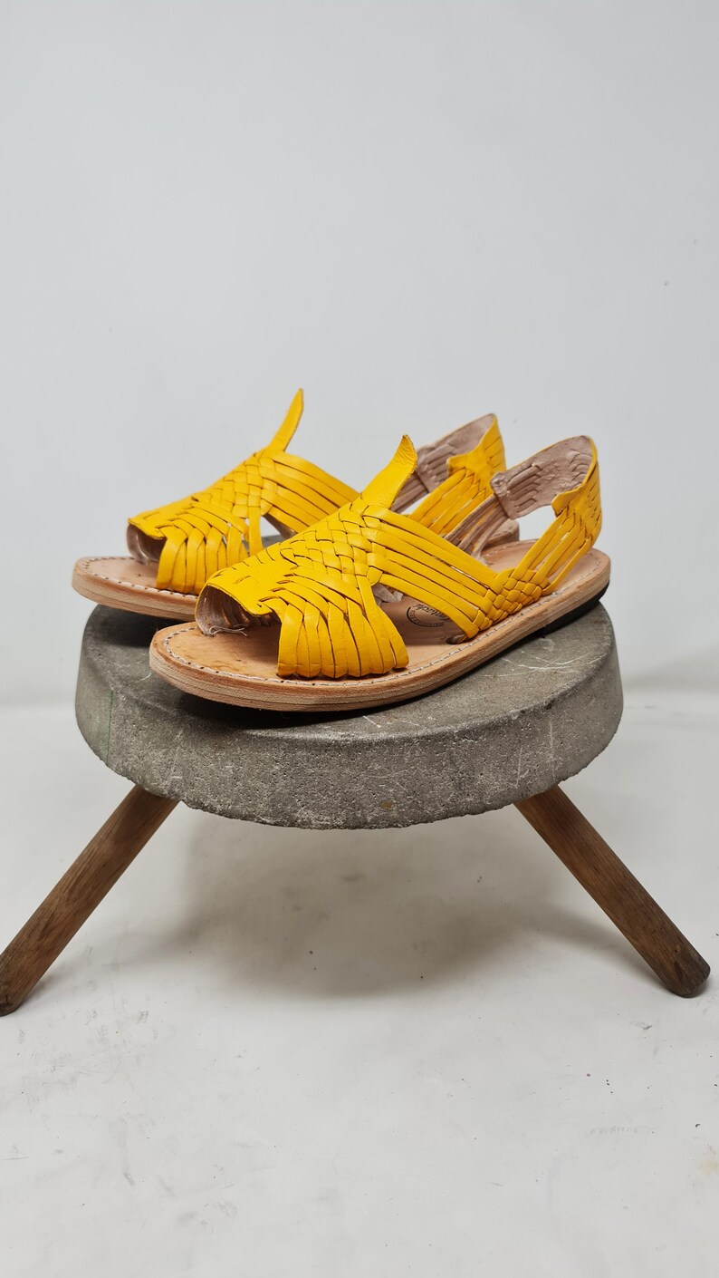 May include: A pair of yellow leather sandals with woven straps. The sandals are sitting on a wooden stool with a stone top.
